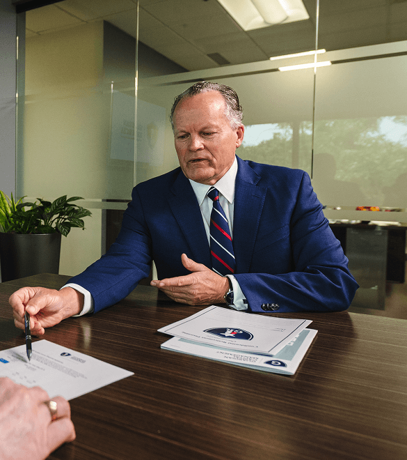 A man in a blue suit and striped tie gestures at a document across a wooden desk in a modern office setting. Another hand holds a pen, poised to sign.