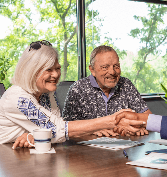 Two older adults sit at a table with documents, smiling and shaking hands with a person across from them in an office with large windows.