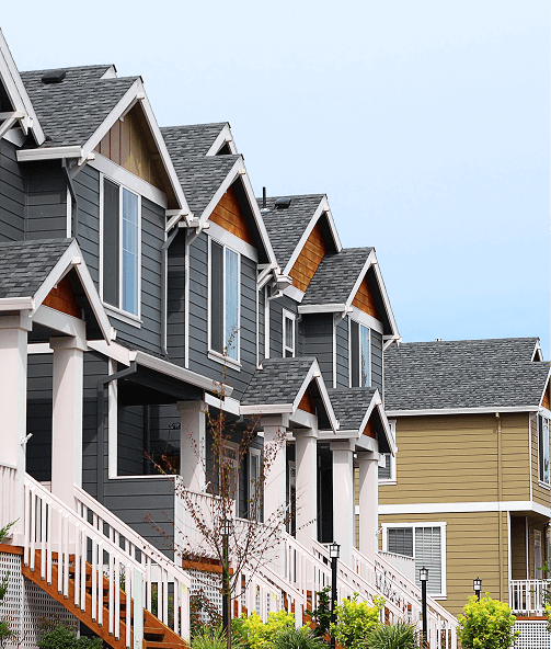 Row of modern suburban houses with grey siding, white trim, and covered porches, set against a partly cloudy sky with landscaping in front.